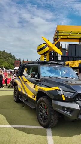 The video shows a black and yellow custom SUV parked on a grassy area. The SUV has a large yellow and black striped orb with a propeller-like structure on its roof. There is a large black and yellow tent-like structure in the background with the word "Билайн" (which translates to Beeline, a telecommunications company) written on it in yellow letters. A motorcycle is parked to the right of the SUV, also with black and yellow accents. Several people are visible in the background, some walking around and others standing near the structure. The sky is blue with scattered clouds. The overall scene appears to be an outdoor event or exhibition.