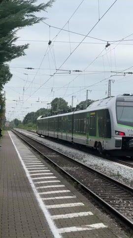 A train is passing through a station in Rees, Germany. The train is silver and green with "VIAS 3" and "ET 25 2301" visible on its front. The train is on a railway track, and there is a platform with white markings next to it. A person is standing on the platform, looking towards the train. The sky is cloudy, suggesting it might be a cooler day. The setting appears to be a rural or suburban area, with trees visible in the background. The overall mood is calm and observant, capturing the moment a train passes by a station.