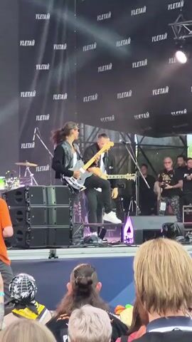 The video shows a live music performance on an outdoor stage. Two musicians are prominently featured in the foreground. The musician on the left is playing an electric guitar and is seated on a speaker stack. They are wearing sunglasses, a black jacket over a white shirt, and black pants with white sneakers. The musician on the right is standing and playing an acoustic guitar. They are wearing a black jacket over a white shirt and black pants. Behind them, several other people are visible on stage, including some wearing t-shirts and hats. The stage backdrop is a large black banner with repeating white text that appears to be "YLETAЙ." The audience is visible in the foreground, with the backs of their heads turned towards the stage. The lighting suggests it is daytime.