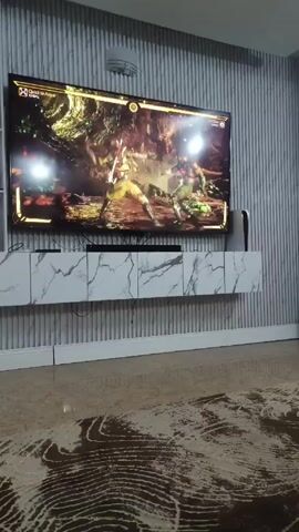 The video shows a TV playing a fighting game, presumably Mortal Kombat, on a modern marble-effect TV stand. The TV is mounted on a wall with vertical stripes, and the floor has a brown patterned rug. No people are visible, only the game being played on the screen. In the background, one can make out a small speaker and what might be a PlayStation 5. The camera appears to be stationary.