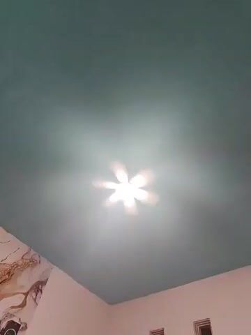 The video shows a ceiling with a light fixture in the center. The ceiling is painted a teal color and has a marble-patterned wallpaper on the left side. The light fixture appears to be a fan with lights, and it is illuminated, casting a glow on the ceiling. The video is filmed from a low angle, looking up at the ceiling. There are also glimpses of two rectangular objects near the bottom of the frame, which appear to be ceiling vents or lights. The overall impression is of a room interior.