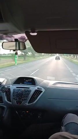 The video shows the interior of a vehicle driving on a road. The dashboard is visible in the foreground, with a radio and air vents. The windshield shows a road ahead with trees and fields on either side. Several other vehicles are on the road in front of the car. The sun is low in the sky, casting a warm light.