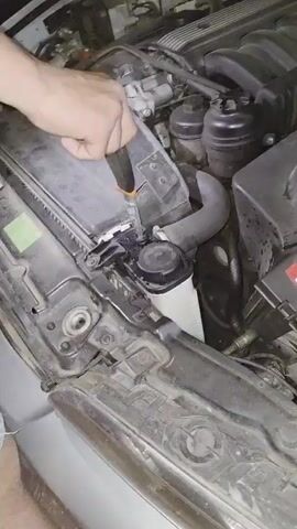 The video shows a person using a screwdriver to open the air filter box of a car. Steam is escaping from the engine compartment. The car is silver and appears to be an older model based on the design of the engine components. The environment is dark, suggesting the video was taken at night. The person is wearing blue jeans and is positioned in front of the open hood of the car, focusing on the engine. The engine bay seems dusty, with some residue and wear visible on the components.