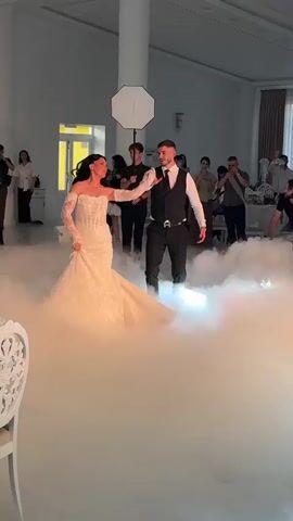 A wedding reception is captured in a dimly lit ballroom. A bride in a white, lace, long-sleeved wedding gown dances with her groom, who is dressed in a black vest and black pants. They are surrounded by a low-lying fog machine creating a hazy, atmospheric effect. Guests stand around the periphery of the dance floor, some holding cameras and phones to capture the moment. The room is spacious with white walls, a large white pillar, and a soft white light illuminating the couple. A white octagon-shaped softbox diffuser hangs from the ceiling. A white ornate chair is visible in the foreground.