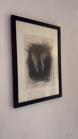 The video shows a framed charcoal drawing of hands hanging on a textured white wall. To the right of the drawing, a window with textured glass is visible. A modern wall sconce with two upward-facing lights is mounted on the wall above the drawing. The scene appears to be indoors, possibly in a home or art studio. There are no people visible in the video.