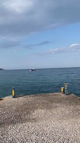 The video shows a wide shot of a calm, blue sea under a partly cloudy sky. A small, white boat with a canopy is seen further out on the water, carrying what appear to be several people. The foreground initially shows a pebbly beach, then transitions to a concrete pier with yellow and blue posts.