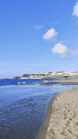 A vertical video shows a calm ocean with gentle waves lapping against a sandy beach. In the distance, buildings line the coastline under a bright blue sky with a few scattered clouds. The water is a deep blue, transitioning to lighter shades as it approaches the shore. The sand appears dark, possibly volcanic. The camera is positioned low, giving a close-up view of the water's surface and the edge of the beach. No people are visible in the frame.