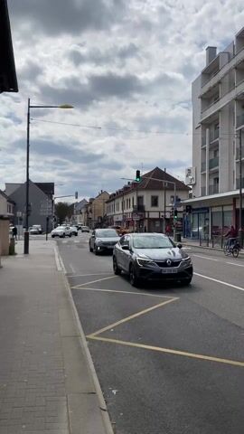 The camera captures a view of a street scene in a European town or city under a partly cloudy sky. A sidewalk paved with grey square stones runs along the left side of the frame, bordered by a street with yellow traffic lines. Cars of various makes and models are visible driving through an intersection where the traffic lights are changing. A person on a bicycle is moving along the street. Buildings of varying heights and architectural styles line both sides of the street, including a taller building with a pale facade and several floors. Street lights and overhead wires are also visible. The camera remains static throughout the short video.
