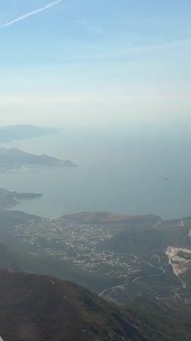 An aerial view captures a coastal landscape. A vast expanse of calm, light blue water stretches across the horizon. In the mid-ground, a settlement with numerous buildings is nestled along the coastline and spreads inland. Winding roads are visible throughout the town and extend up the sides of the rocky, green hills that dominate the foreground. A significant construction or mining site with exposed earth and roads is prominent on one of the hills. A few dark-hulled ships are discernible in the water. The sky is a pale blue, with a thin, white contrail visible in the upper left quadrant. The overall impression is one of a developing area situated between mountains and the sea.