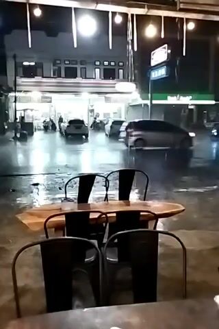 The scene is set at night in a commercial area, possibly a town square, viewed from what appears to be an outdoor dining area. Heavy rain is falling, creating reflective surfaces on the pavement and the street. In the foreground are two sets of metal chairs with wooden seats and backs, arranged around a wooden table.  Across the wet street, there are several buildings.  One is a two-story building with numerous windows and lit interiors.  Next to it is a green-colored commercial establishment with lit signage in what appears to be Chinese characters.  Several vehicles, including cars and a motorcycle, are moving through the rain-soaked streets, kicking up sprays of water.  The overall lighting is provided by a combination of streetlights and the lights emanating from the buildings.