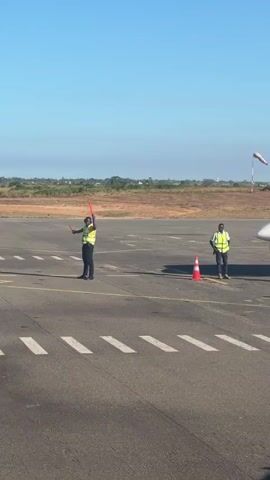 The video shows an airport tarmac in Pemba, Mozambique, on a clear, sunny day. Two ground crew members, wearing yellow reflective vests, are standing on the asphalt. The crew member on the left is holding red marshalling wands and giving hand signals, indicating activity related to an aircraft. Another crew member is positioned further to the right, near an orange traffic cone. A third individual, wearing a white shirt, dark pants, and a reflective vest, briefly walks into the frame from the right, interacts with the second ground crew member, and then moves towards the aircraft. The nose and part of the wing of a white aircraft are visible on the far right. In the background, a wind sock is visible, and the landscape appears flat with dry ground. The sound of an aircraft engine is audible throughout the clip. A partial aircraft registration, starting with "V", can be seen on the side of the plane.