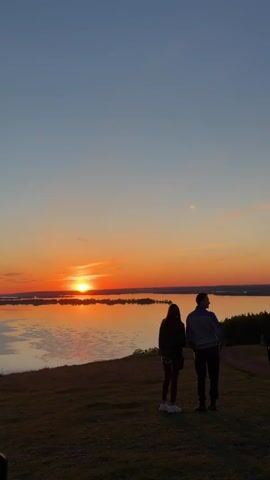 A medium vertical shot depicts a couple silhouetted against a vibrant sunset, standing on a grassy knoll overlooking a body of water. The sun is setting directly in front of them, casting a bright orange glow over the water and sky, with the sky transitioning from orange near the horizon to a clear blue above. The couple is standing side by side, the woman on the left, slightly shorter than the man. The woman has long dark hair and is wearing a dark jacket and a short skirt. The man is wearing a jacket and dark pants. They are both facing the sunset. To the right, a treeline is visible along the edge of the water. Other people are vaguely visible in the background to the right. The woman raises her hand, as if to shield her eyes from the sun, then puts her arm around the man's shoulder.