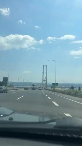 The video shows a car driving on a highway toward a large suspension bridge. Several other cars and a bus are also traveling on the highway. The sky is blue with white clouds, and there are hills in the distance. The bridge has two tall towers and multiple suspension cables. Streetlights line both sides of the highway. The camera is positioned from inside the car, looking through the windshield. There is a green sign beside the highway. A person is speaking in the background.