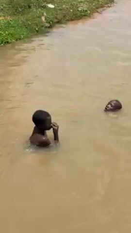 The video shows two children swimming in a body of muddy water. The water appears to be shallow enough that both children can stand. One child is closer to the camera, partially submerged. The second child is slightly farther away, with only their head visible above the water. The water is brown and murky. Green vegetation is visible along the bank of the water.