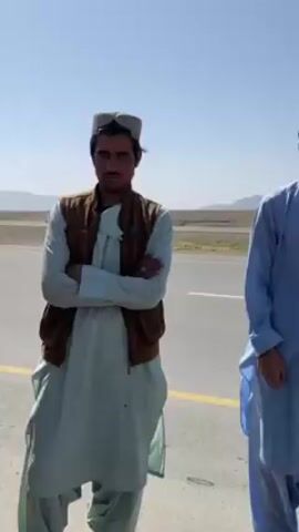 The video shows a scene with four young men standing beside a road on a sunny day. The road stretches out into a desert landscape with mountains in the background. Initially, two of the men are in the foreground, wearing traditional clothing, including hats and long shirts. One man is wearing a brown vest. The camera then moves to the side, showing a man with a black beanie and scarf, and finally to another man with a black outfit. The men appear to be interacting or possibly posing for the camera.