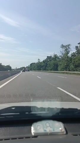 The video shows a first-person view from inside a car driving on a highway. The highway has multiple lanes, and there are other vehicles on the road, including trucks and cars. The weather is clear and sunny, with a blue sky and scattered clouds. Lush green trees line the side of the highway. In the foreground, part of the car's dashboard is visible, including a small religious icon. The camera is focused on the road ahead.