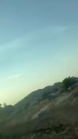 The video shows a view from inside a vehicle moving along a road. The landscape outside is slightly hilly with dry vegetation. In the distance, there is a small settlement with simple buildings. The sky is light blue, and the sun is shining brightly, causing glare. The camera appears to be pointed upwards and slightly tilted. A tree is visible in the foreground.
