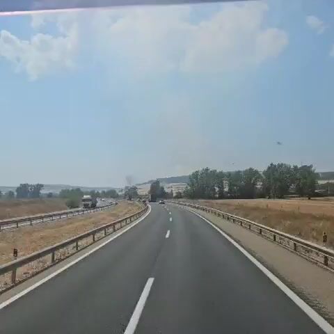 The video shows a POV shot from a vehicle driving on a highway on a clear, sunny day. The highway has multiple lanes, separated by white lines and a metal guardrail. The landscape is rural, with dry, golden fields and scattered trees on either side. In the distance, on the left side of the highway, there is a plume of smoke rising into the sky, suggesting a fire. Several vehicles, including trucks and cars, are visible on the highway, moving in the same direction as the camera. The trees in the foreground on the right are green and lush, contrasting with the dry fields. The sky is bright blue with a few scattered clouds. The overall impression is one of travel through a rural area, with the unusual element of smoke visible in the distance.