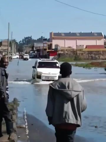 The video shows a flooded street. A white car drives through the water, creating waves.  Two young men stand on the sidewalk watching the car. A building with the words "KALIMBURU INTEGRATED ACADEMY" is visible in the background. Other cars can be seen farther down the flooded street. The sky is clear.