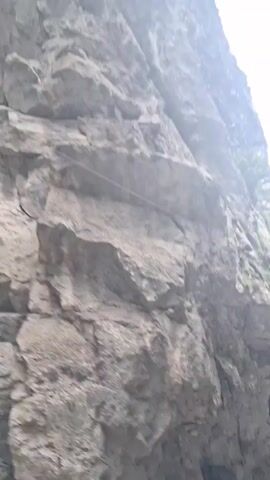 The video captures a scenic view of a road passing through a mountainous area with steep, rocky cliffs on one side and a dense forest on the other. The road appears to be paved and relatively narrow, with a guardrail visible in some parts. A few vehicles, including cars and what looks like a truck, are parked on the side of the road. Several people are seen walking and standing near the vehicles, some of whom appear to be tourists. The overall impression is of a natural landscape that is accessible to visitors, with signs of human activity present. The sky is overcast, suggesting a cloudy day. The camera is moving, indicating that the video was likely shot from a moving vehicle.