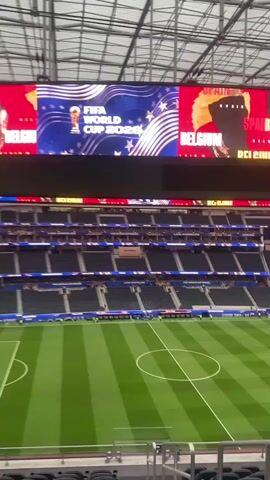 The video shows a panoramic view of a large, empty stadium from the upper seating area. The stadium features a green soccer field with white markings. Above the field, a massive video screen displays graphics related to the FIFA World Cup 2026. The screen is divided into sections, with one side showing the FIFA World Cup trophy and the text "FIFA WORLD CUP 2026" against a blue background with stars and wavy lines. The other side of the screen shows the flags of Spain and Belgium, with the word "BELGIUM" visible. The seating areas around the stadium are filled with empty, dark-colored chairs. The stadium has a modern, retractable roof structure. In the distance on the field, several individuals wearing yellow vests are visible, along with some soccer goals. The overall atmosphere is quiet and devoid of a crowd.