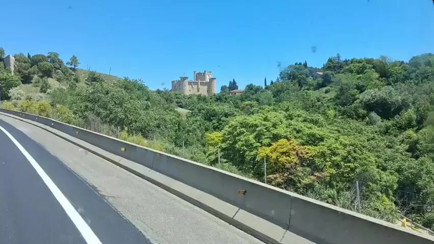 The video shows a stone castle on a hill overlooking a highway. The camera appears to be inside a vehicle moving along the highway. The castle is surrounded by lush green trees and bushes. The sky is clear and blue. The camera quickly zooms in on the castle before becoming obscured by the surrounding trees.