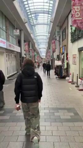 The video shows a person walking through a covered shopping arcade. The arcade has a high, glass-paneled roof allowing natural light to filter through. Numerous shops line both sides of the walkway. Red and pink decorative banners hang overhead. Several people are walking in both directions. The person recording the video is walking forward, and their back is visible. They are wearing a black puffer jacket and camouflage pants. Another person in a similar black jacket and gray pants is walking slightly ahead and to the left. Some of the shops have awnings or signs in Japanese. The floor is paved with gray bricks. A clothing rack is displayed outside one of the shops.