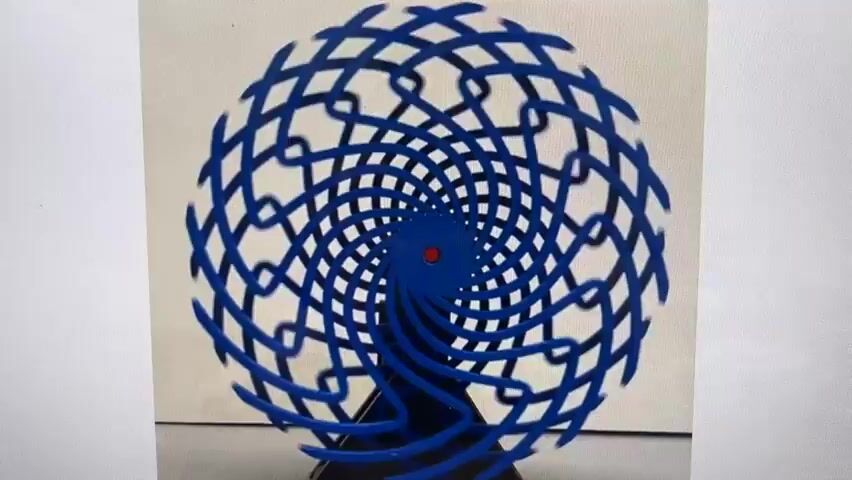 A blue geometric sculpture is displayed against a white background. The sculpture has a spiraling, latticework design that extends outwards from a central red dot. The sculpture appears to be in motion, with some parts blurred due to the movement. The base of the sculpture is also blue and has a multi-layered, curved shape. The lighting is even, suggesting an indoor setting.