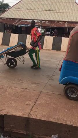 The video captures an outdoor street scene in Isieke, Nigeria. Three individuals are visible. A man wearing a pink and red short-sleeved shirt with green pants pushes a loaded metal wheelbarrow along a paved road, moving from right to left across the frame. The wheelbarrow carries items covered by a blue tarp.

Adjacent to the road, a concrete sidewalk is separated from a deep, open gutter filled with debris and trash. On the sidewalk, another man in a light-colored shirt and shorts bends down to handle several large white sacks. A third person, partially visible in a patterned dress, stands near these sacks.

The paved road is bordered by concrete barriers painted with black and white stripes. In the background, various buildings with different architectural styles and colors, including one with a red roof and another with green and orange accents, line the street. Utility poles and lines are also visible. A sign in the background displays the text "CATON JOHN BEST NEST".

The soft, diffused lighting suggests either early morning or late afternoon under a clear sky. No specific emotions are discernible from the individuals.