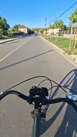 The video shows a first-person perspective of someone riding a bicycle on a paved road. The rider's hands are visible on the handlebars, which have a phone mount attached. The road appears to be in a rural or suburban area, with houses, trees, and utility poles visible on either side. The sky is clear and blue, and the sun is shining, casting long shadows. The rider is moving forward, and the scenery is passing by.