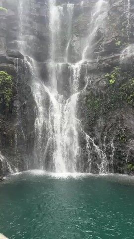 A vertical video shows a waterfall cascading down a rocky cliff face into a pool of water below. The rocks are covered in green moss and vegetation. The water from the waterfall creates a mist that obscures the top of the cliff. In the background, there is a wooden walkway with two people walking on it. The walkway is surrounded by lush green trees and foliage. The video is filmed from a low angle, looking up at the waterfall.