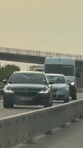 The video shows a series of cars and trucks driving on a highway under an overpass. The sky is overcast, and the lighting suggests it is either dawn or dusk. The vehicles are driving in traffic, with some cars following closely behind each other. The first car seen is a silver Volkswagen Golf, followed by a white Mercedes-Benz Sprinter van. Behind the van is a red truck. Then, another white Mercedes-Benz Sprinter van with black racing stripes is visible, followed by a black car. Finally, a red truck is seen with a black car behind it. The overpass above the highway has a metal railing.