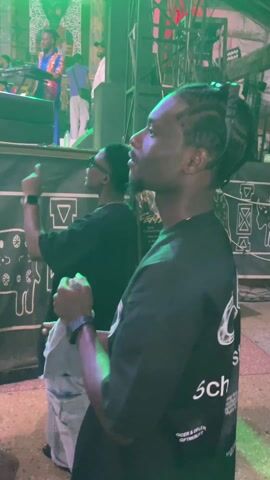 Here's a description of the video:
The video captures a live music performance in a dimly lit indoor venue, likely a concert or event space. The focus shifts between a man in the foreground and the band performing on stage. The man in the foreground wears a black t-shirt with a white design, light-colored patterned pants, and a watch. He holds a light-colored cloth in his hands and appears to be enjoying the music, occasionally gesturing with his fingers. Another man in a black t-shirt stands behind him.

The camera then pans up to show the stage. The band consists of multiple members playing instruments such as keyboards, guitars, and drums. The lead singer is shirtless, wearing a blue wrap-around skirt. Behind the band, large banners with images and the name "SHAKARA" are visible. The stage is bathed in vibrant green and blue lighting, and spotlights shine down on the performers. A few other audience members are visible in the foreground, with their heads and shoulders partly obscuring the view of the stage.