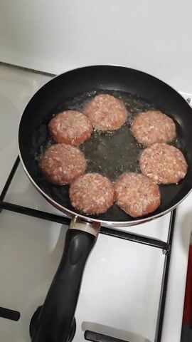 Here's a description of the video: 

A hand places raw, round patties of what appears to be ground meat into a black frying pan on a white stovetop. Seven similar patties are already in the pan, sizzling in a generous amount of oil. The patties are light pink with dark flecks and are spaced around the pan, not touching each other. The stove burner is lit beneath the pan.