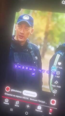 The video displays a screenshot from a tablet device, showing a portion of a video playing. In the foreground, a man of East Asian descent wearing a blue baseball cap and a tactical vest is visible. The cap has a circular emblem with an abstract design. The vest is dark and appears to have multiple pockets. Text overlays are present on the screen. One overlay in Russian reads "@kinolatsay" followed by "Подписаться" (Subscribe). Below this, more Russian text says "Отсылка на сериал MEOW. Фильм Патрул..." (Reference to the series MEOW. Film Patrol...). Another overlay in Russian, written in a glowing purple font, appears to be dialogue or captions: "Алмате был ме" and "вместе задчиков кощ марили". The background is blurred but suggests an outdoor, possibly wooded, setting with trees and foliage. On the right side of the screen, there are interface elements, including a heart icon, a share icon, and a "..." (more) icon, along with numbers indicating likes and views. The bottom of the screen shows a navigation bar typical of a video platform, with icons for Home, Shorts, a plus sign, Subscriptions, and Profile.