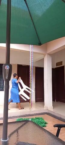 A woman in a blue dress is carrying a white chair into a building with an "Eye Clinic" sign. The video is shot from under a green patio umbrella, showing a colorful plastic chain hanging from the umbrella. A table with artificial grass decor sits nearby. The woman walks through an open doorway into the building.
