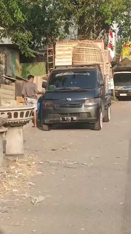 A black minivan is parked on a dirt road, with a person standing next to it. The minivan has two large wooden cages stacked on its roof. Two decorative concrete planters are on the side of the road. In the background, there are trees, buildings, and piles of trash. A red metal gate is in front of a closed shop.