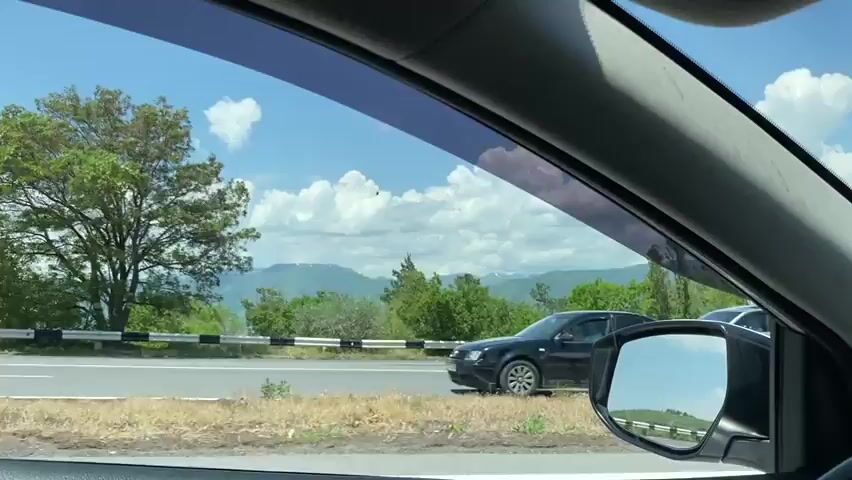 A first-person view from inside a car shows the road ahead. The sky is blue with scattered white clouds. Green trees line the side of the road, with some taller, slender trees standing out. A mountain range is visible in the distance. The car's side mirror reflects another vehicle behind. The interior of the car is visible, including the dashboard, steering wheel, and sun visor. The car appears to be on a highway.