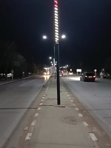 The video shows a night view of a street with cars driving on it. On both sides of the street, there are sidewalks. Between the sidewalk and the road, there is a median with a tall, black light pole. The light pole has white lights spiraling around it and red and green lights at the top. There are trees on the left side of the street. There are streetlights above the road. The sky is dark.