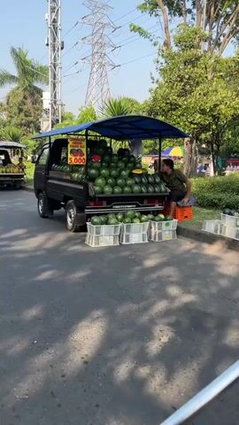 The video shows a street lined with fruit vendors selling from the back of pickup trucks. The first truck in focus is black and heavily laden with watermelons, with some stacked in plastic crates in front. A sign on the truck advertises watermelons for 5,000. A person in a light-colored hijab stands near the back of the truck. In the background, other trucks are parked, some selling different fruits, possibly durian, and a few people are seen interacting with the vendors. There are trees and a tall power pylon in the background. A few motorcycles are also visible on the street. The scene appears to be a busy outdoor market.