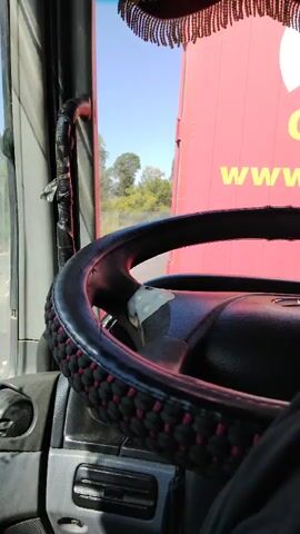 This is a first-person perspective video from inside a truck cab, looking out the driver's side window and towards the steering wheel. The video shows the road and passing scenery through the window, with reflections in the side mirrors. The steering wheel is covered in a decorative fabric. The overall environment appears to be a sunny day on a highway. The truck appears to be in motion.