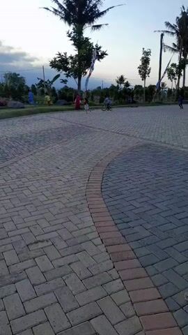 A video shows a paved area with interlocking bricks, with palm trees and other greenery in the background. The sky is cloudy, suggesting dusk or dawn. Several people are visible: two children are riding bicycles, and one person appears to be running or walking. In the foreground, a motorcycle's rearview mirror is visible, reflecting a person's hand. The scene appears to be a public park or recreational area.