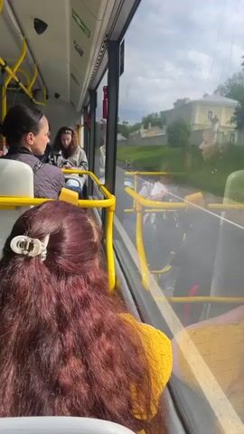 The video shows the interior of a bus with several passengers. In the foreground, a person with long, dark red curly hair and a yellow top is seated, facing away from the camera, looking out the window. To their left and further down the aisle, a woman with dark hair pulled back is seated and looking at her phone. Behind her, another woman with curly brown hair is also seated and looking at her phone. Further down the bus, several other passengers are visible, some standing and some seated. The bus has yellow handrails and poles. Outside the window, greenery and buildings are visible, suggesting the bus is in motion. The lighting indicates it is daytime.
