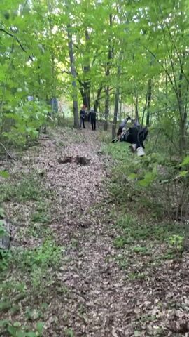 The video shows a wooded area with a steep, leaf-covered slope. At the top of the slope, a group of people are gathered around a person who appears to be lying on the ground. Some of the people are wearing helmets and protective gear, suggesting they may be engaged in an outdoor activity such as zip-lining or adventure sports. There is a zip line cable visible running across the frame. One person is assisting the individual on the ground, while others observe. The environment is dense with trees and undergrowth, indicating a natural, undeveloped setting.