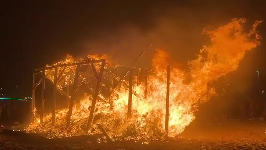 This media file captures a large bonfire on a sandy beach at night. The central subject is a wooden structure, resembling a hut or a small building, engulfed in flames. Intense orange and yellow fire billows from the structure, illuminating the dark sky and casting a warm glow on the surrounding sand. The framework of the burning structure is clearly visible, with some parts collapsing into the inferno. Smoke rises into the night air, obscuring the background in some areas.

In the distance, on the left side of the frame, a few indistinct figures of people can be seen watching the spectacle. There are also faint lights, possibly from vehicles or distant buildings, suggesting the presence of a populated area nearby. On the right side, more indistinct figures are gathered, observing the fire. The overall scene evokes a sense of a communal event or celebration, likely taking place on the beach in La Línea de la Concepción, Spain.

The time of day is clearly night, indicated by the dark sky and the dominance of firelight. The weather appears clear, as there is no visible rain or strong wind affecting the fire's behavior. The sandy beach setting is evident from the ground material. The emotions conveyed are a mix of awe and perhaps excitement, typical of observing a large, controlled fire. No visible text is present in the media file.