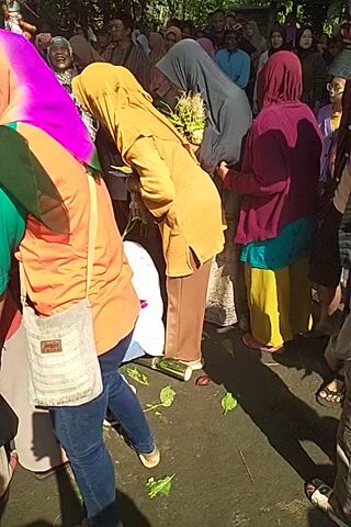 Here is a concise description of the video:

The video shows a group of people outdoors, kneeling and touching or reaching towards a wrapped object on the ground. Most appear to be women wearing headscarves, along with a few men who are also looking down at the scene.  Green leaves are scattered around on the concrete surface. The camera is held at an angle looking down towards the group.
