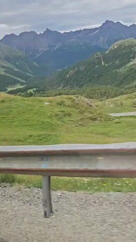 The video shows a scenic mountain landscape. In the foreground, there is a metal guardrail along a road. Beyond the guardrail, rolling green hills covered in grass and some small bushes are visible. Further in the background, the landscape transitions to steeper, wooded mountains with some rocky peaks. A winding road can be seen cutting through the hills. The sky is overcast with clouds. The video appears to be shot from a moving vehicle.