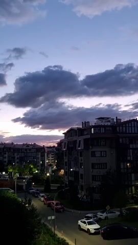 The video begins by capturing a view of a residential area at dusk. Tall buildings with multiple windows and balconies are visible, some with lights on. A street runs through the scene, with several cars parked along its sides and driving on it. The sky is filled with dramatic, dark clouds, with streaks of purple and pink light indicating the setting sun. The overall atmosphere is calm and serene, with the urban landscape set against a beautiful natural phenomenon.