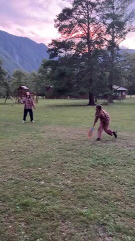 Two people are playing badminton in a grassy field with cabins and trees in the background. The sky is a dusky purple and pink, indicating it's either sunrise or sunset. The person on the left, wearing a checkered shirt and dark pants, is swinging a badminton racket. The person on the right, wearing a brown tracksuit, is also swinging a racket, facing away from the camera. Mountains are visible in the distance. The overall impression is a leisurely outdoor activity in a scenic natural setting.