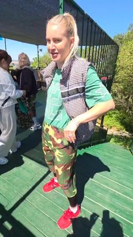 The video, likely filmed in Jiejinkou, China, shows a woman with blonde hair tied in a ponytail, standing on a green wooden deck under bright, sunny daylight. She is wearing a green t-shirt, a houndstooth patterned vest, camouflage shorts over black leggings, and red sneakers. A bright green mesh object is visible, seemingly tucked into the back of her shorts. In the first shot, she smiles and adjusts her shorts. She then turns around, revealing the words "CHRISTIAN DIOR" printed on the back of her vest. Other people are partially visible in the background, along with white buckets and metal railings, suggesting an outdoor setting like a boat or a viewing platform. The overall atmosphere appears light and playful.